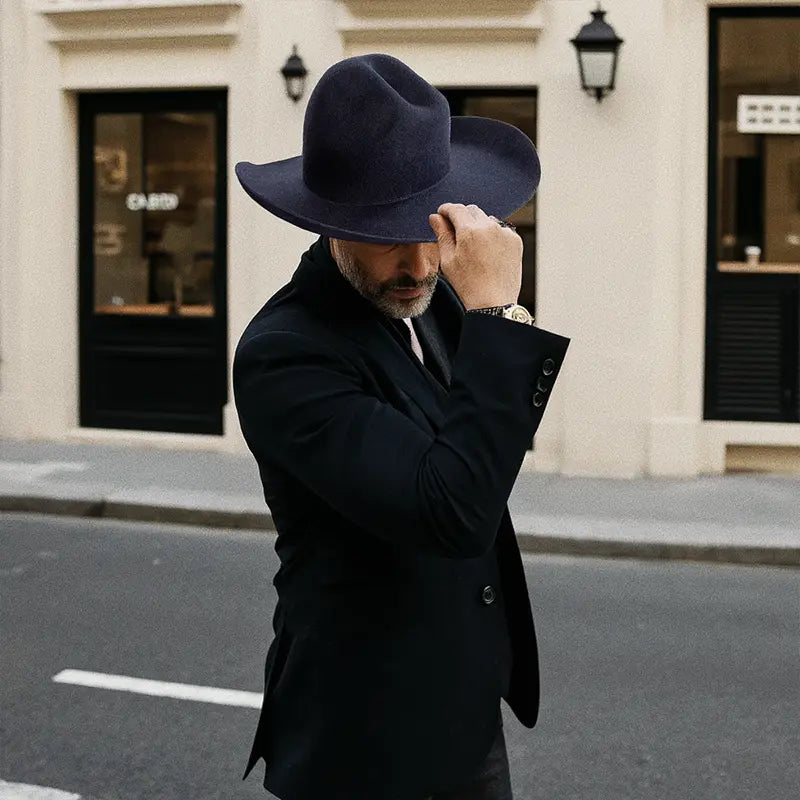 Outside a building with a black awning, a formally dressed man wore a navy blue, wide-brimmed felt cowboy hat.