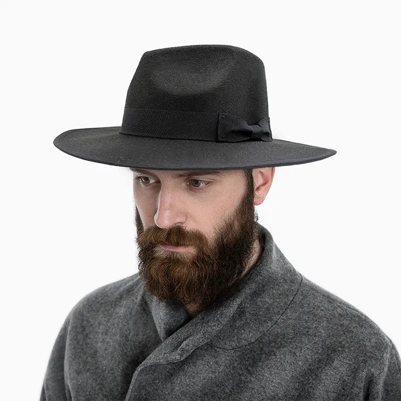 Man wearing a black wide - brimmed hat with a black band and a small bow detail, paired with a gray coat and a full beard